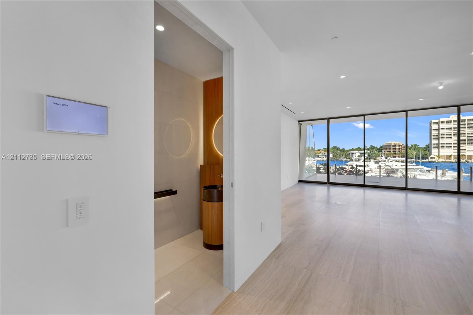 Floor-to-ceiling glass wall, wood-finish flooring, recessed lighting, and wall-mounted smart home control panel, featuring a powder room with cylindrical vanity and circular back-lit mirror Floor-to-ceiling glass wall, wood-finish flooring, recessed lighting, and wall-mounted smart home control panel, featuring a powder room with cylindrical vanity and circular back-lit mirror