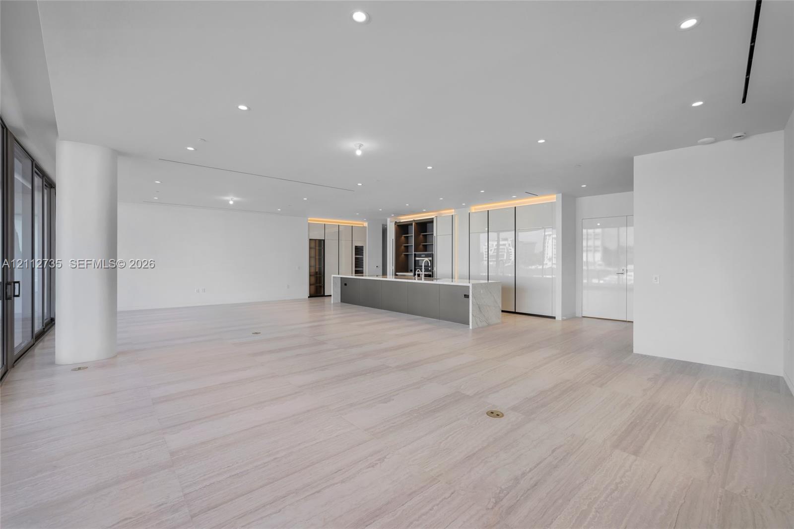 Open-concept space featuring light-toned wood-finish flooring, recessed ceiling lighting, floor-to-ceiling glass sliding doors, and a kitchen island with a stone-finish countertop and integrated cabinetry