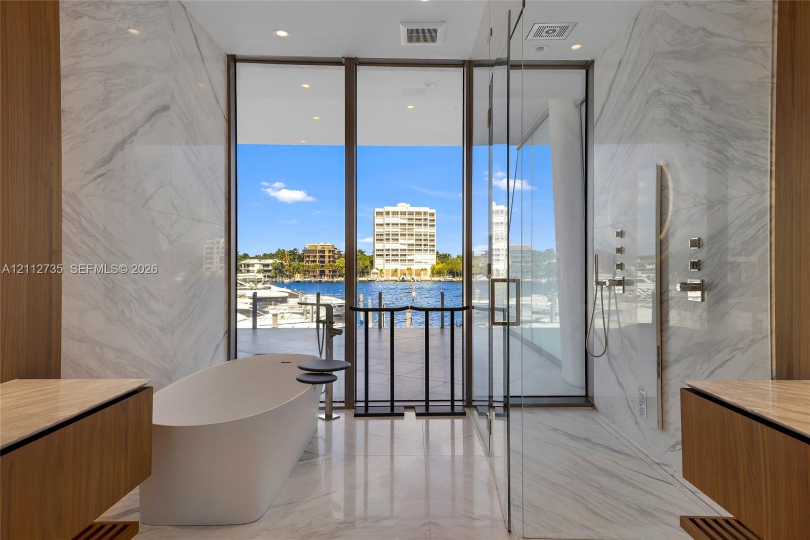 Floor-to-ceiling glass windows, stone-finish wall cladding, freestanding soaking tub, glass-enclosed shower, and dual wood-finish floating vanities