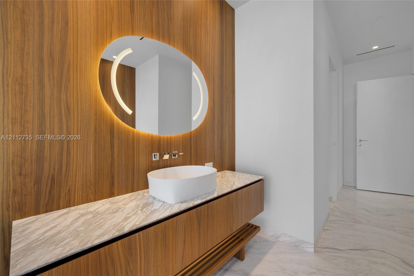 Floating wood-finish vanity with a stone countertop, white vessel sink, wall-mounted faucet, and oval backlit mirror set against a floor-to-ceiling wood-paneled wall