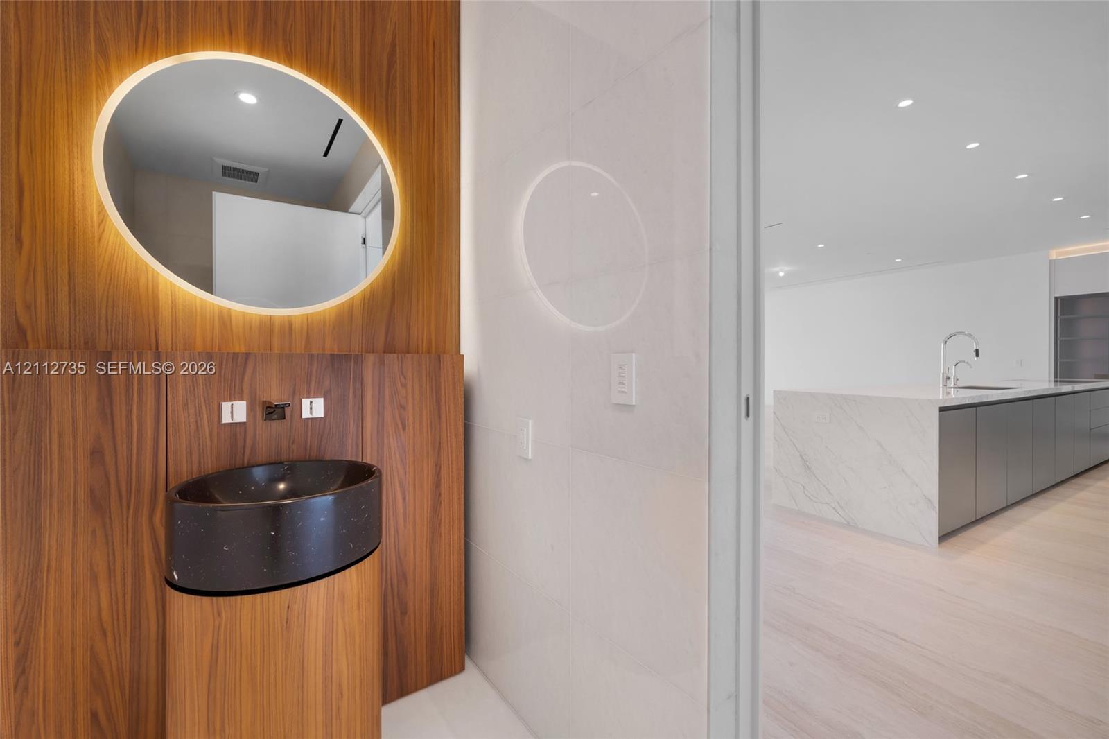 Wood-paneled wall feature with integrated backlit oval mirror, dark speckled pedestal basin, wall-mounted faucet, and stone-surfaced kitchen island with matte cabinetry Wood-paneled wall feature with integrated backlit oval mirror, dark speckled pedestal basin, wall-mounted faucet, and stone-surfaced kitchen island with matte cabinetry