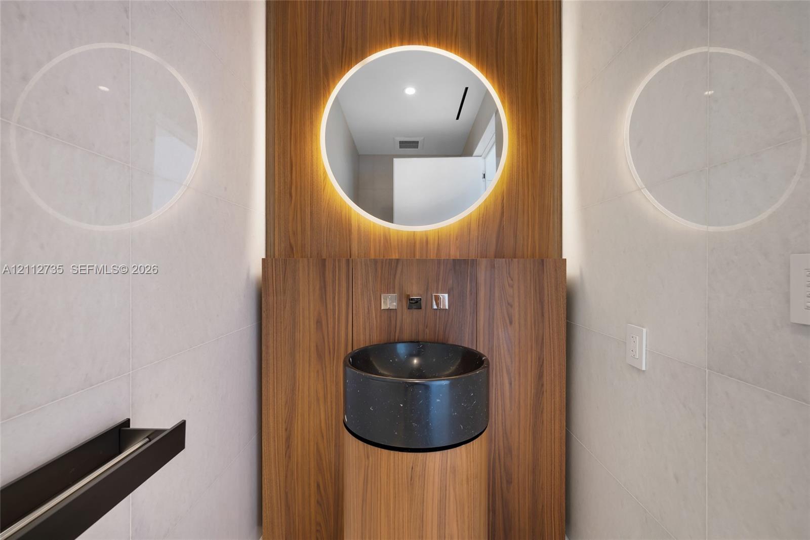 Wall-mounted wood-finish paneling, circular backlit mirror, cylindrical wood-finish pedestal vanity, black stone vessel sink, and wall-mounted metallic fixtures Wall-mounted wood-finish paneling, circular backlit mirror, cylindrical wood-finish pedestal vanity, black stone vessel sink, and wall-mounted metallic fixtures