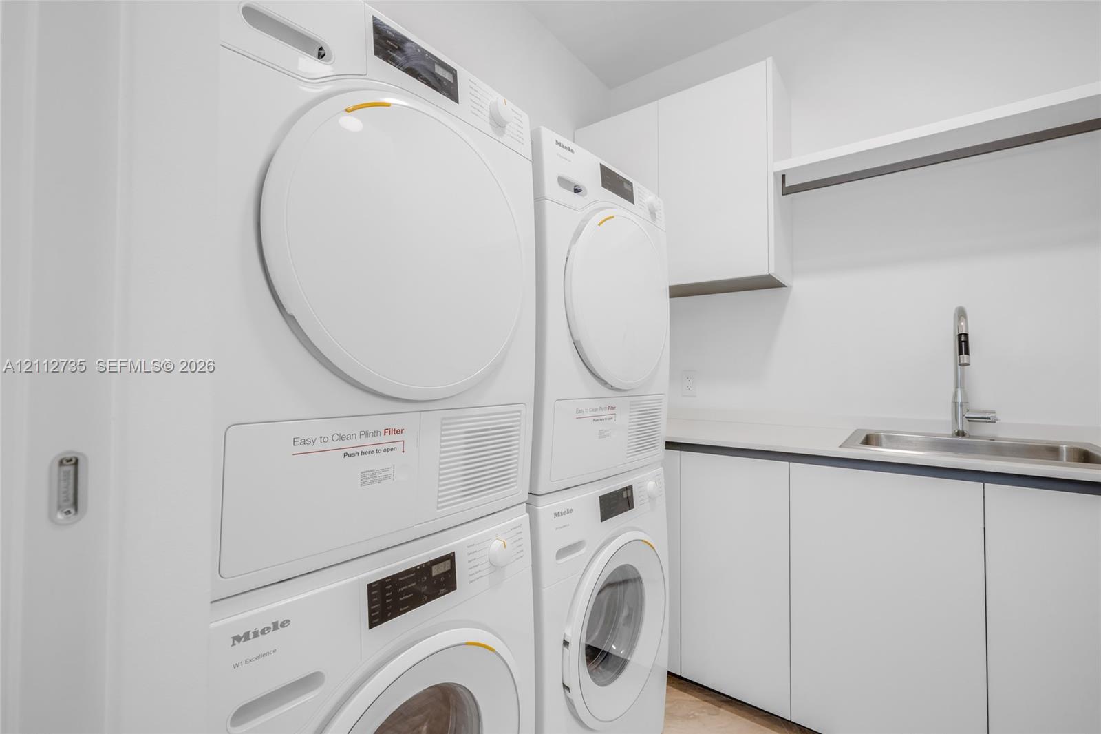 Laundry room featuring stacked front-load appliances, built-in cabinetry with solid-surface countertops, an undermount sink, and wall-mounted shelving