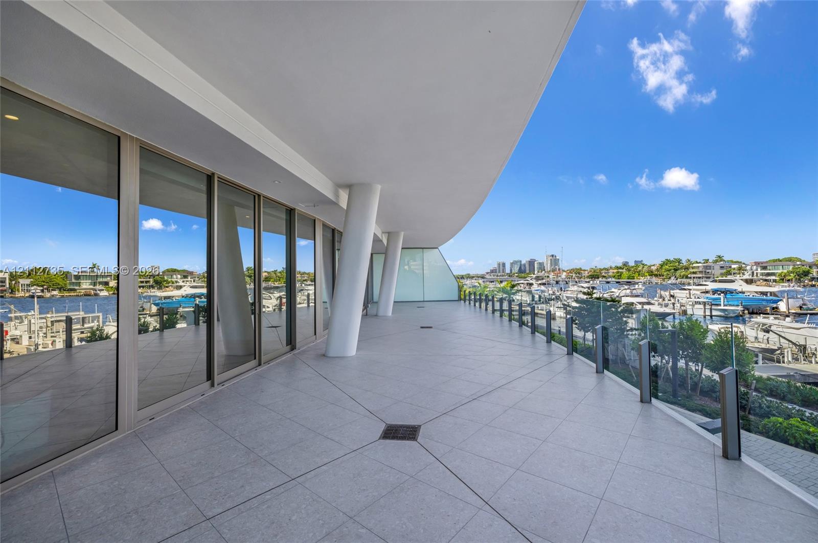 Expansive covered terrace featuring large-format tile flooring, structural support columns, floor-to-ceiling glass wall panels, and a frameless glass railing