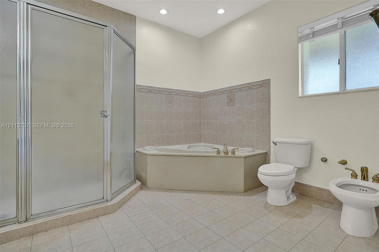 Corner soaking tub with tile surround, glass-enclosed shower, porcelain toilet, and bidet featuring metallic hardware