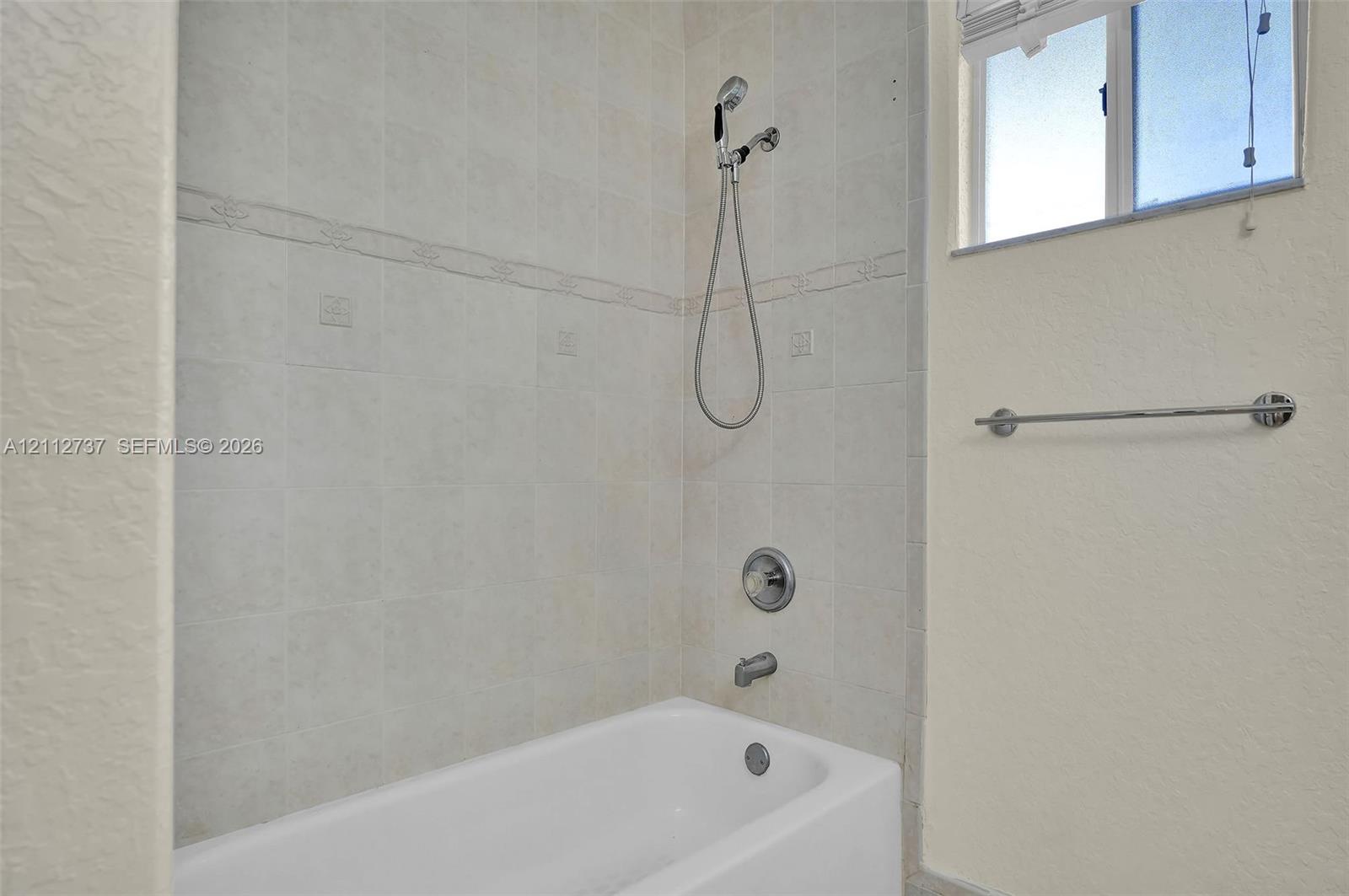 Bathtub and shower combination featuring neutral ceramic wall tile, a handheld showerhead, a chrome faucet assembly, and an integrated towel bar