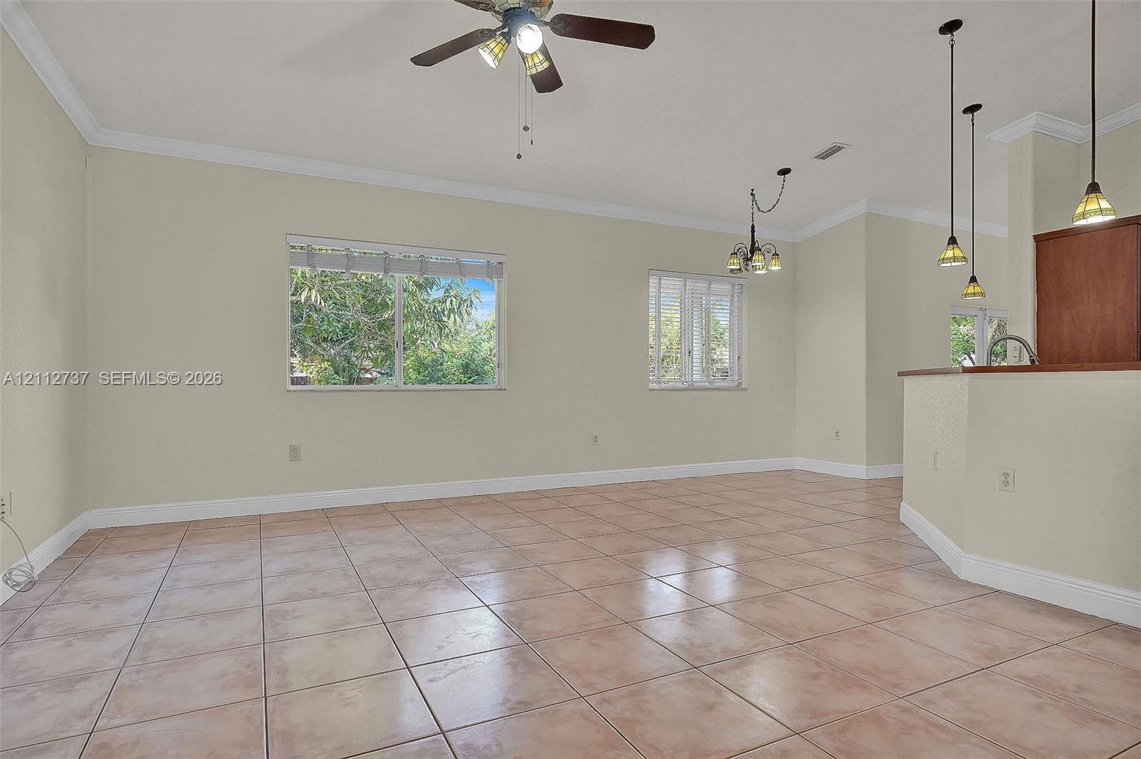 Open living area featuring large-format ceramic tile flooring, crown molding, a central ceiling fan with integrated lighting, and multiple pendant lights over a breakfast bar