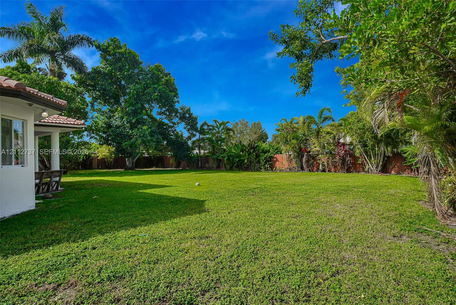 Expansive backyard featuring a large grass lawn, perimeter wood fencing, and mature tropical landscaping