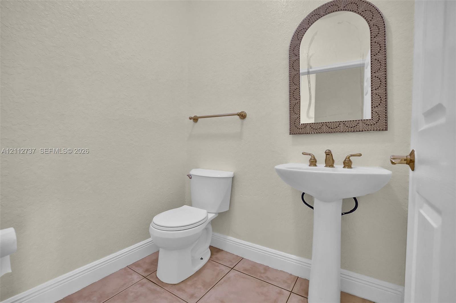 Pedestal sink with gold-tone hardware, arched wall mirror with decorative frame, neutral wall finishes, and tiled flooring