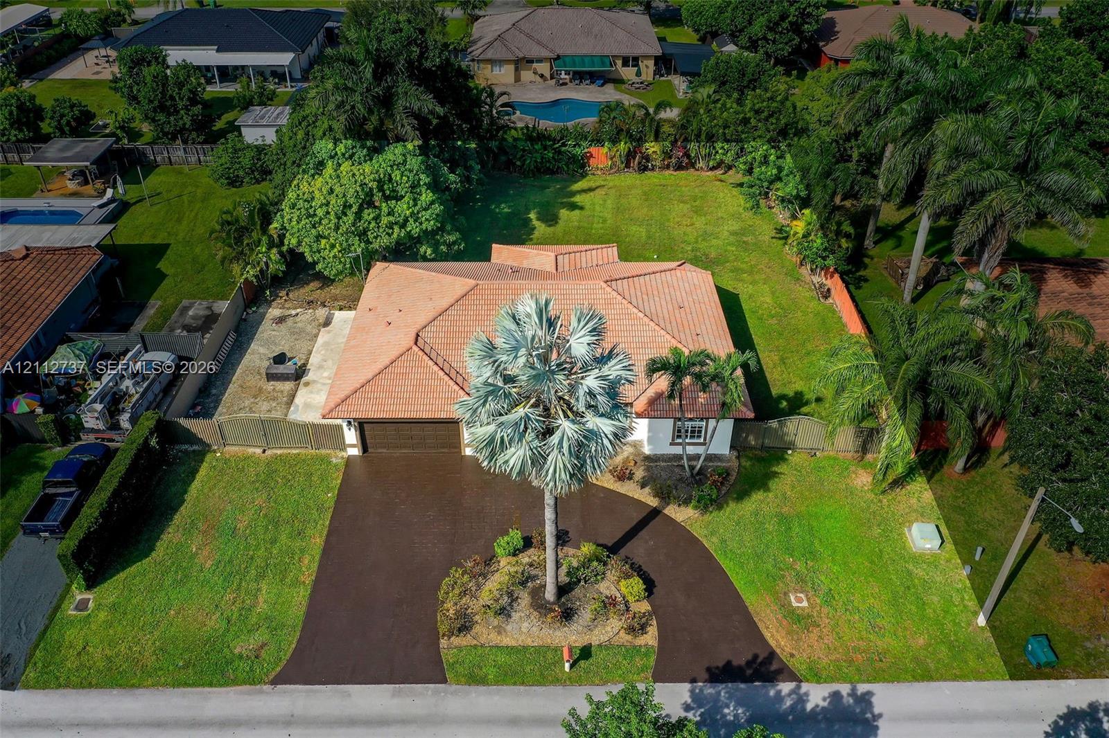 Aerial perspective of a property featuring a multi-hipped tile roof, a circular asphalt driveway, and expansive lawn space Aerial perspective of a property featuring a multi-hipped tile roof, a circular asphalt driveway, and expansive lawn space