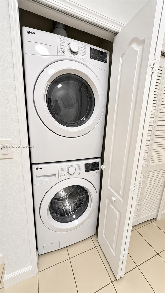 Stacked front-loading washer and dryer unit enclosed in a closet featuring white louvered doors and ceramic tile flooring
