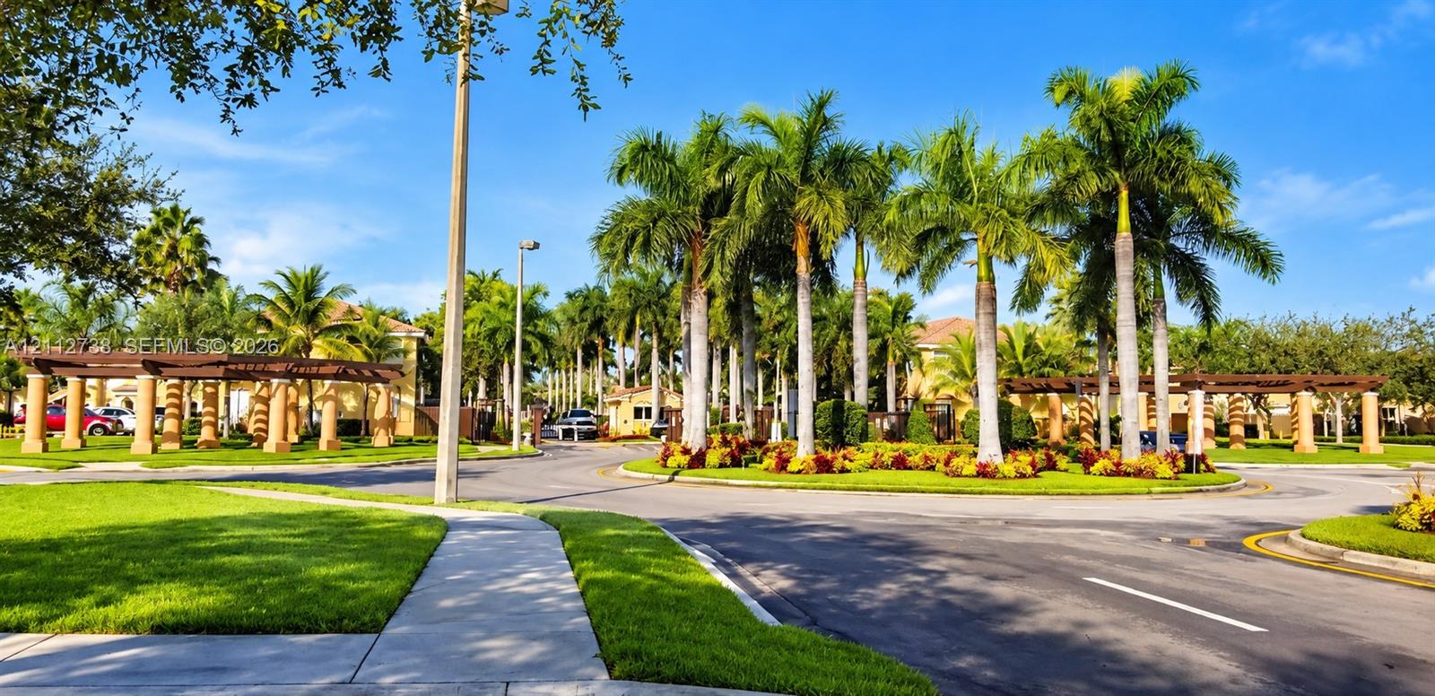 Landscaped entrance featuring mature palm trees, decorative pergola structures, concrete sidewalks, and manicured green lawns