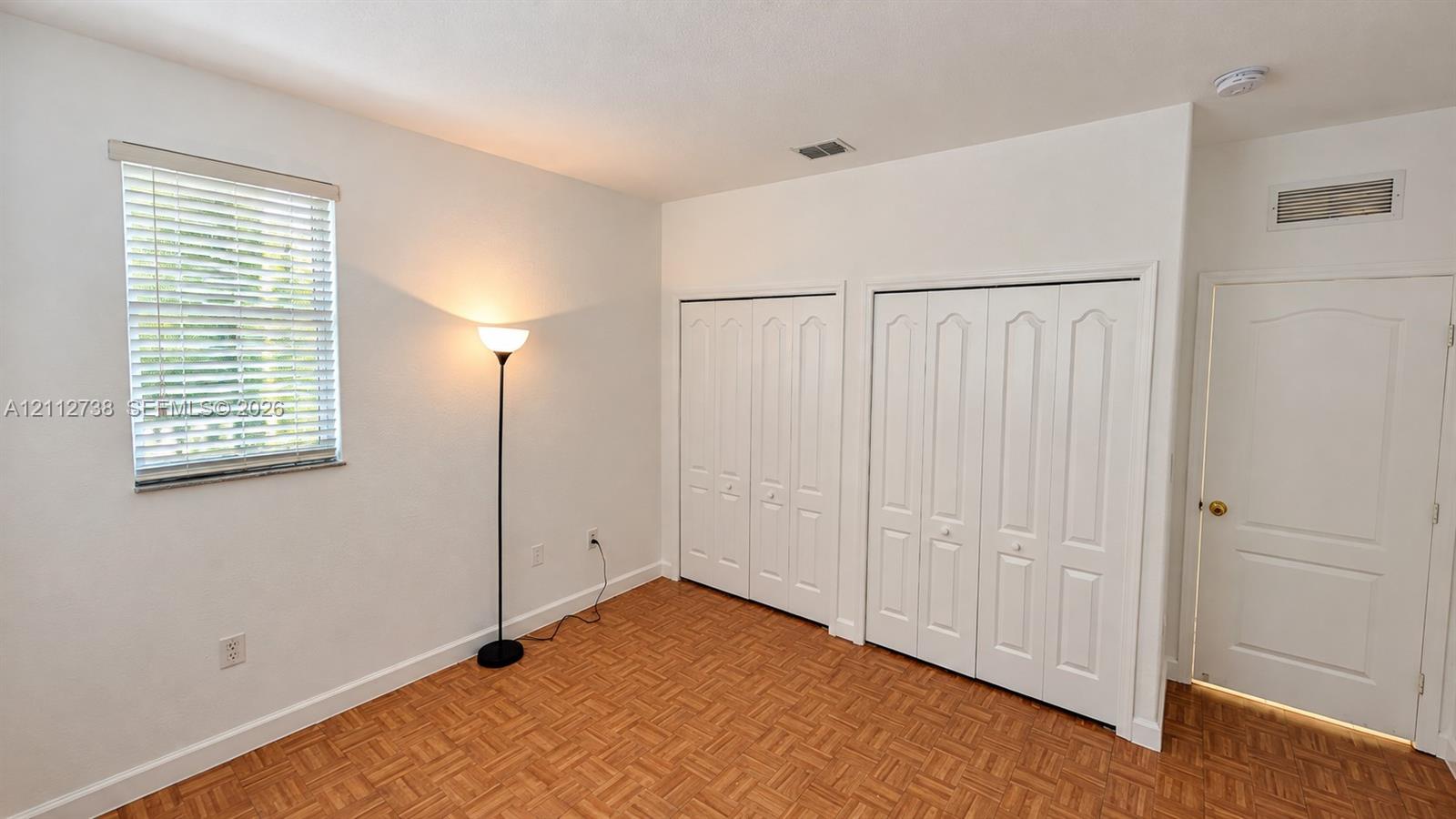 Room with wood-finish parquet flooring, window with horizontal blinds, and two sets of white bi-fold closet doors
