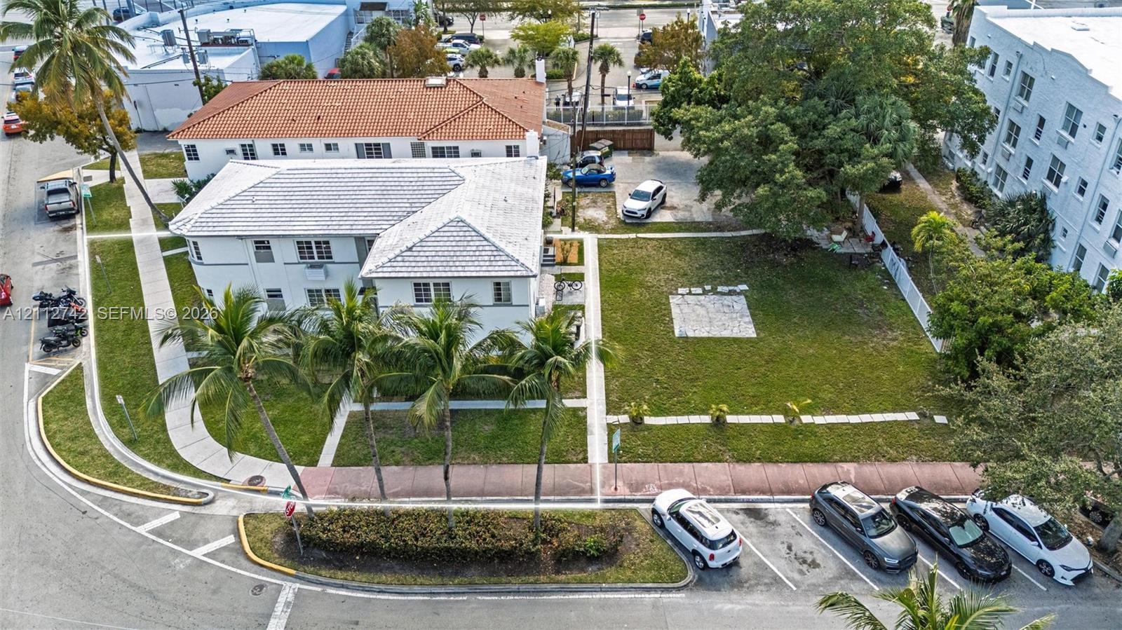 Corner-lot property featuring a multi-story exterior with a light-colored finish, tiled roof, mature palm trees, expansive lawn space, and concrete walkways
