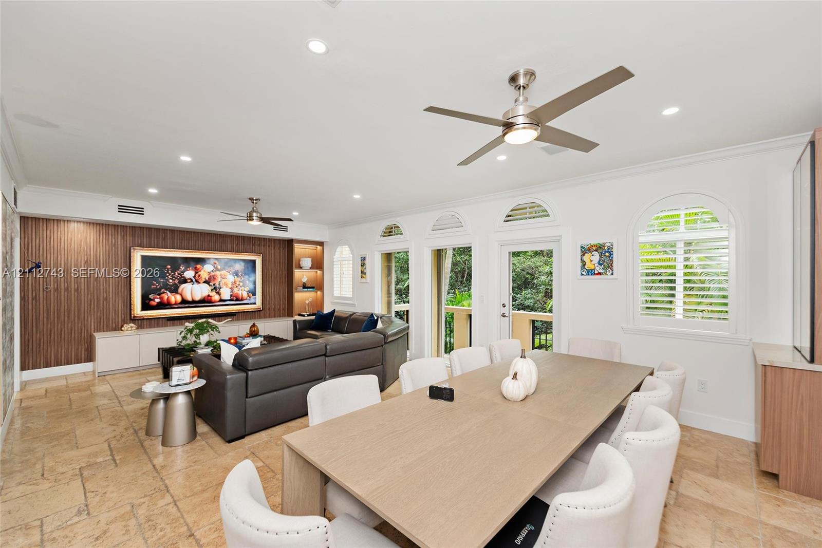 Living area featuring tile flooring, wood-finish wall paneling with integrated shelving, recessed lighting, ceiling fans, and arched windows with plantation shutters