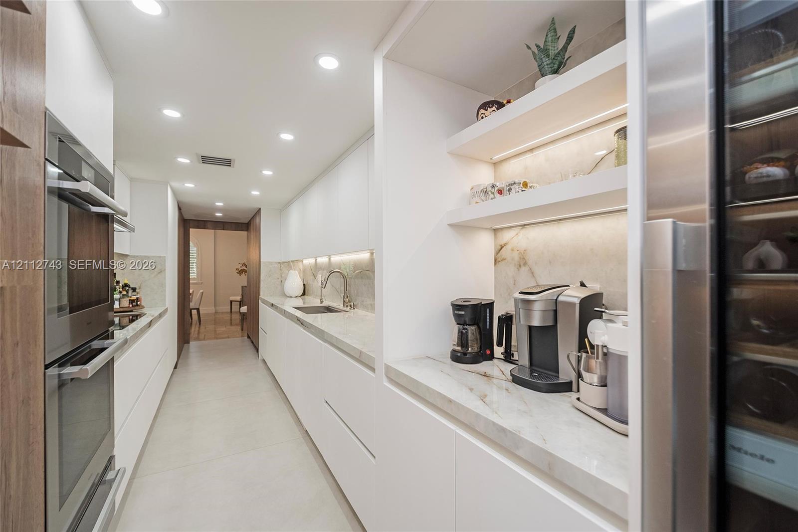 Sleek kitchen featuring flat-panel cabinetry, stone countertops, integrated stainless steel double wall ovens, built-in recessed shelving with under-cabinet lighting, and a stainless steel wine refrigerator