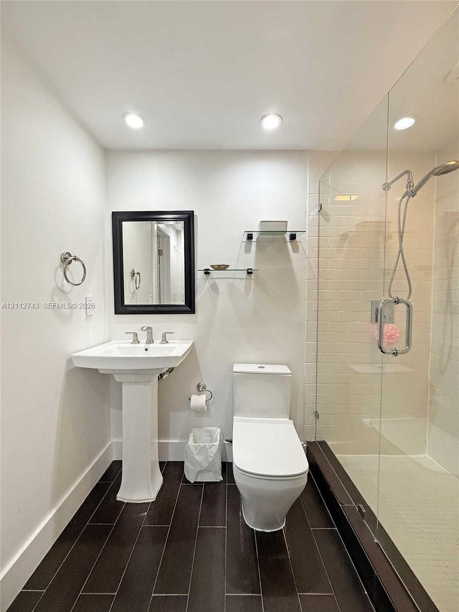 Pedestal sink with chrome widespread faucet, walk-in shower featuring a glass enclosure and wall-mounted hand shower, dark tile flooring, and integrated glass shelving