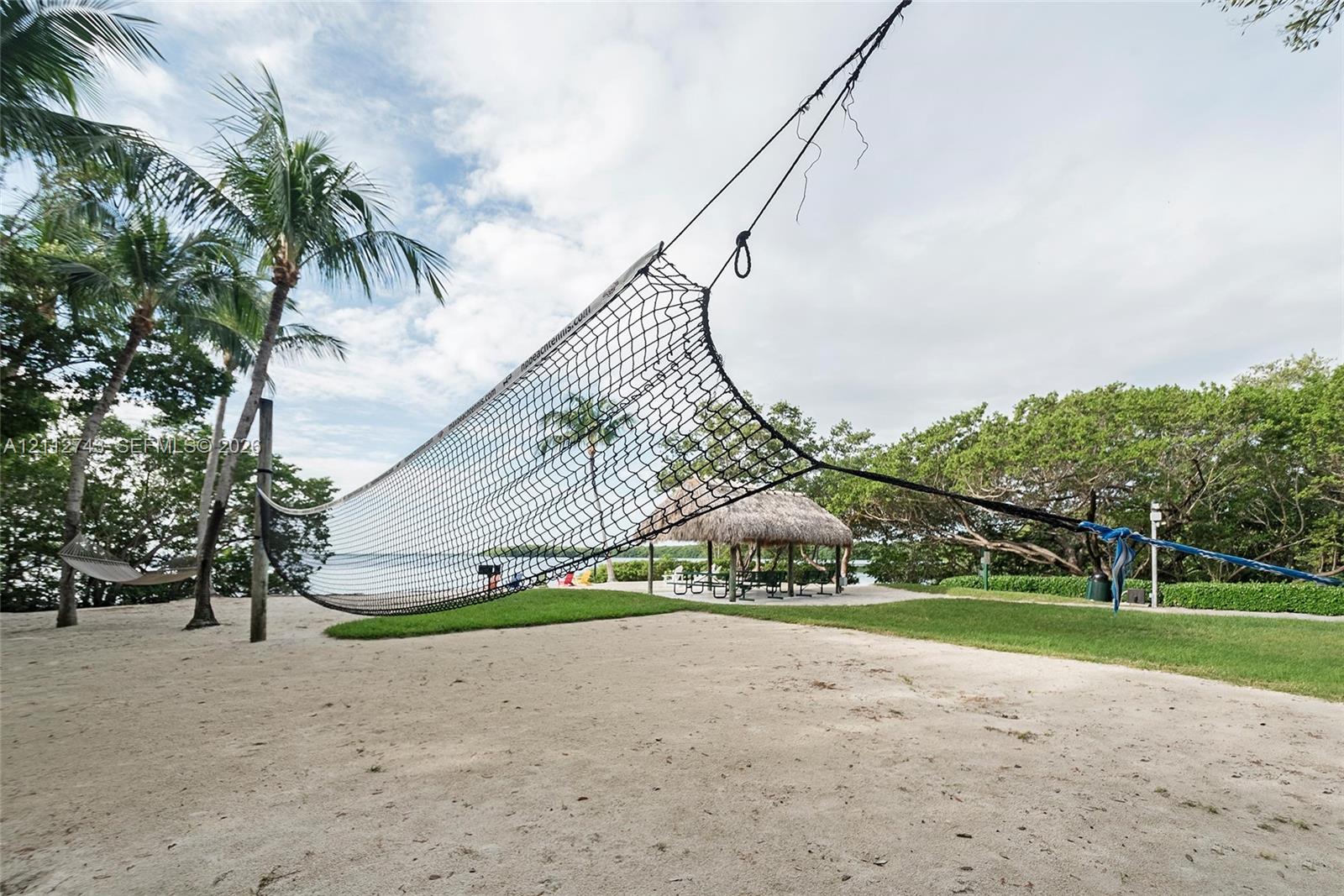 Sandy outdoor recreation area featuring a volleyball net, grass lawn, thatch-roofed gazebo, and mature palm trees