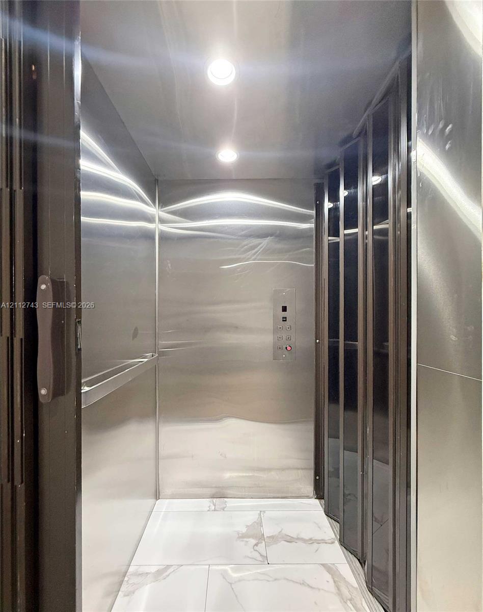 Private elevator featuring polished stainless steel wall panels, recessed ceiling lighting, a horizontal handrail, and large-format tile flooring with a veined pattern