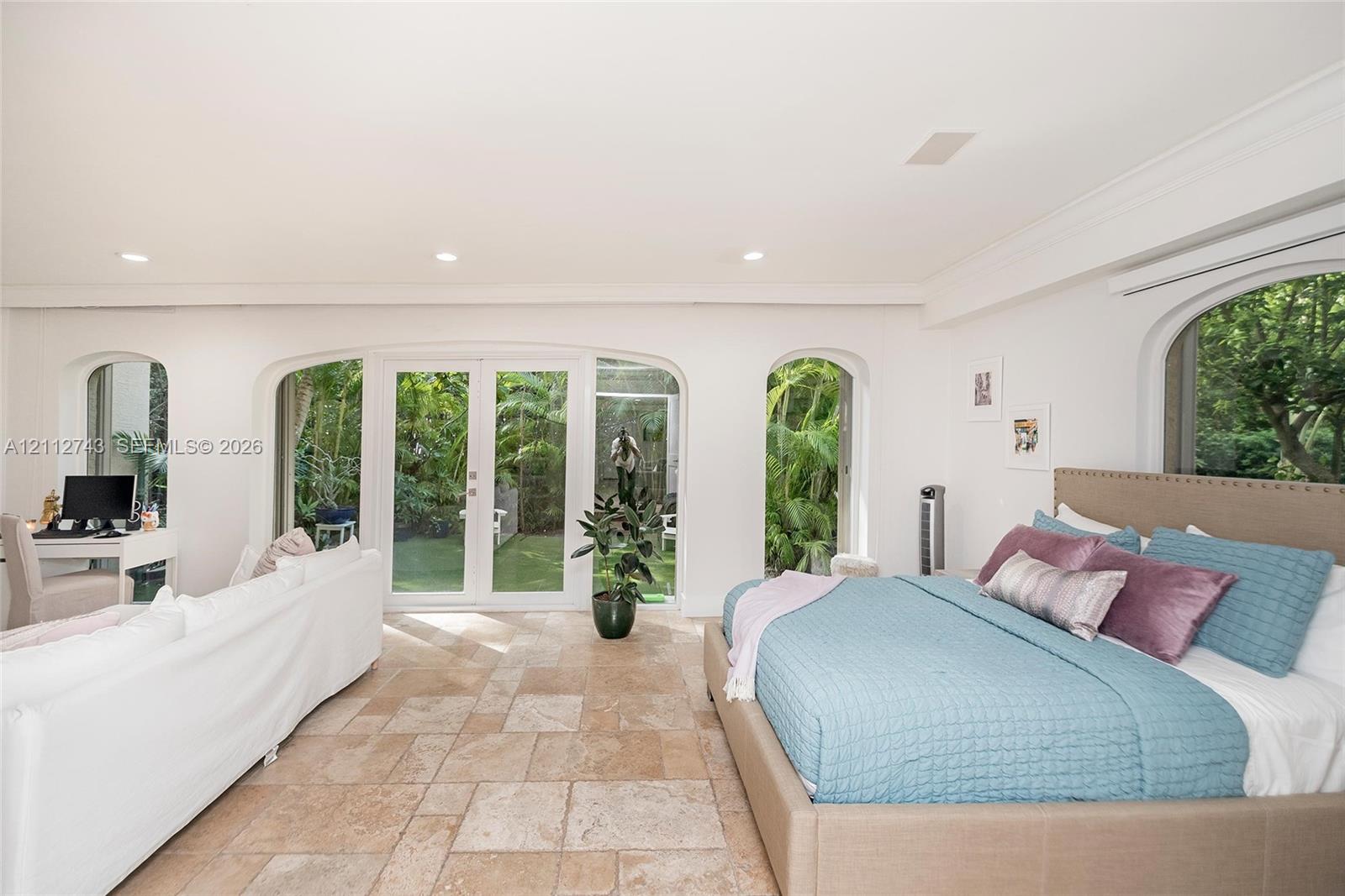 Spacious room featuring light stone-finish tile flooring, arched window casings, crown molding, and glass-paneled double doors providing outdoor access