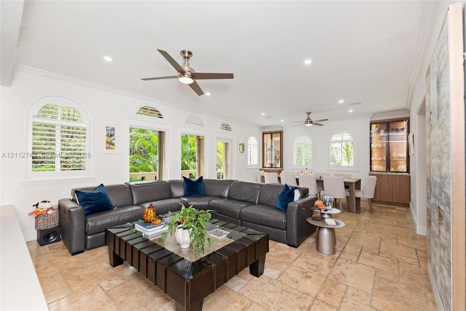 Open-concept interior featuring large-format tile flooring, multiple arched windows with built-in shutters, dual ceiling fans, and integrated custom cabinetry