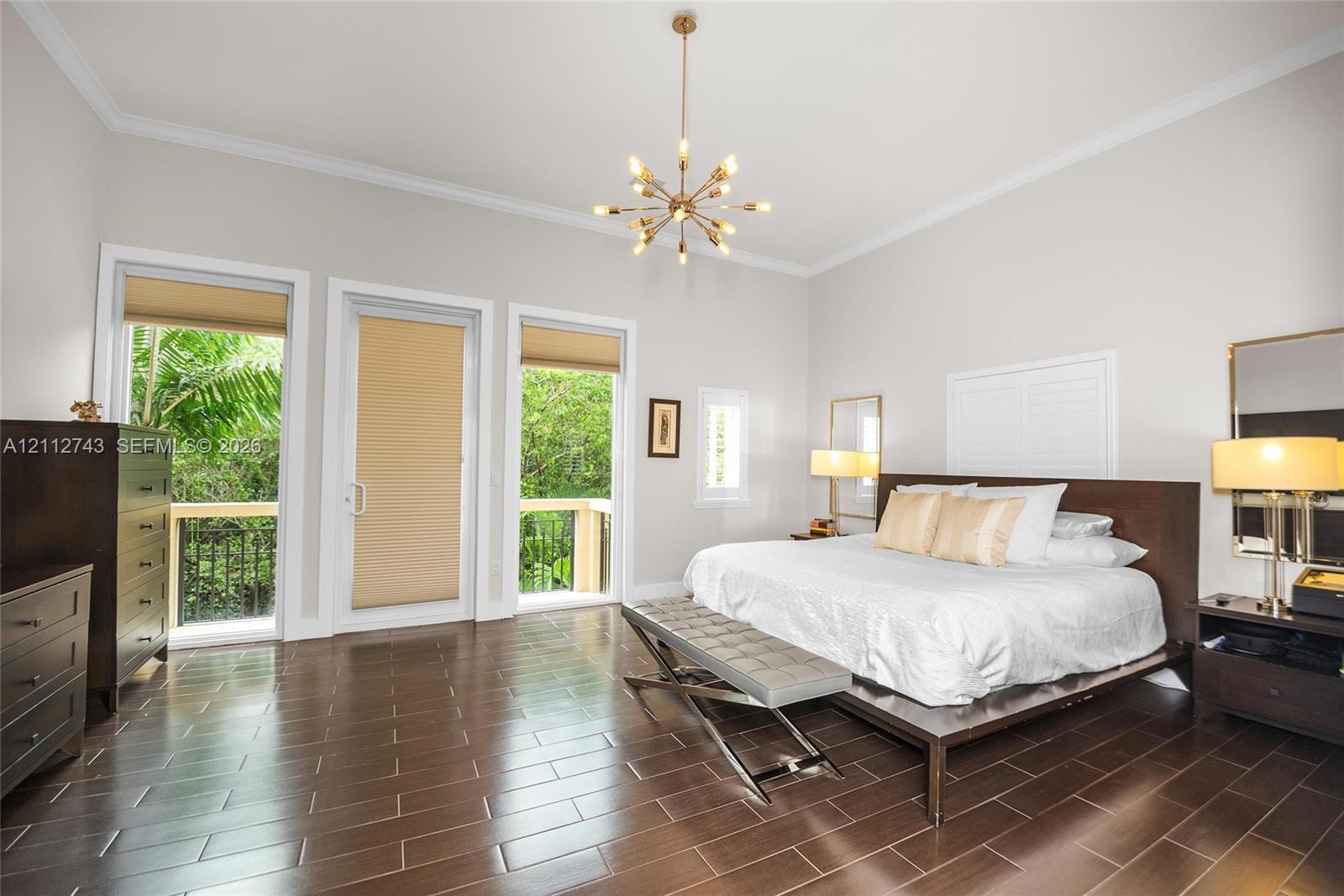 Spacious room featuring wood-finish tile flooring, crown molding, a central multi-arm chandelier, and two glass-panel doors leading to an exterior balcony