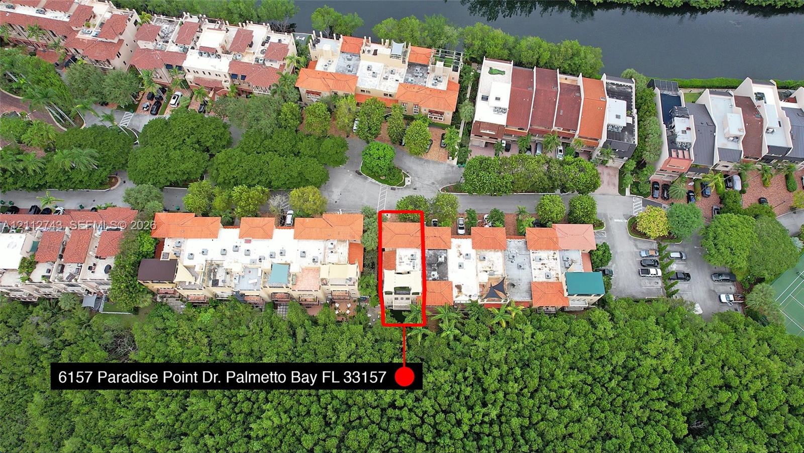 Aerial perspective of property featuring a multi-story structure with a terra cotta tile roof, positioned on a waterfront lot with dense perimeter landscaping