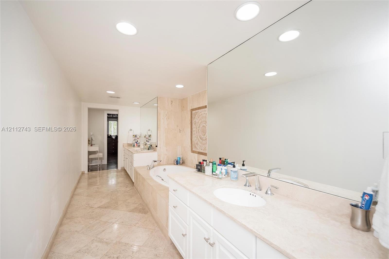 Bathroom featuring stone-finish flooring, a jetted soaking tub with a tiled surround, a lengthy stone-surface vanity with dual integrated sinks, and expansive wall-to-wall mirrors