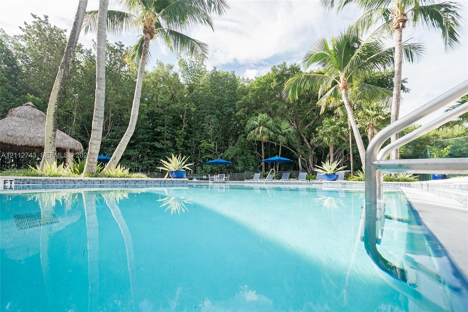 In-ground swimming pool featuring a tiled waterline, metal handrails, a thatched-roof cabana, and mature palm trees