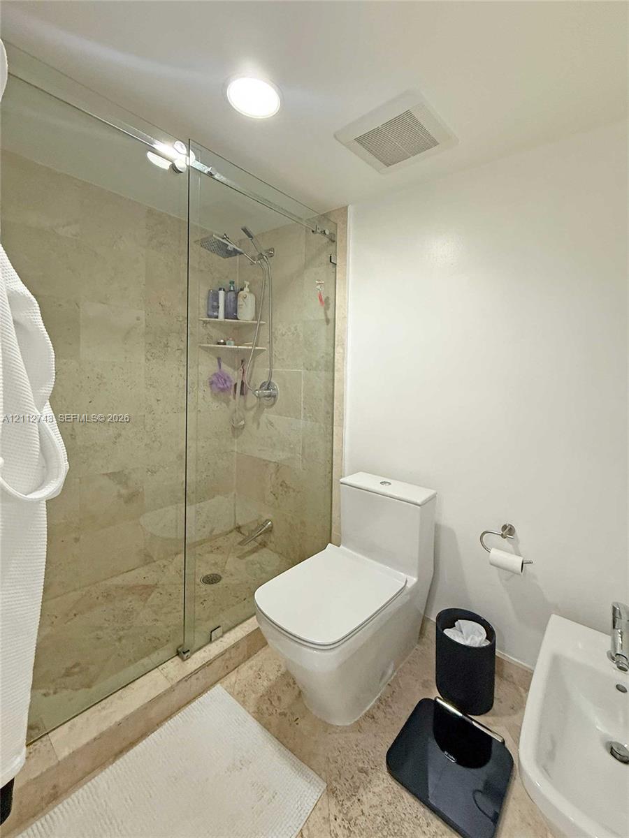 Walk-in shower featuring a frameless glass enclosure, stone tile surround, and a handheld shower fixture, complemented by natural stone flooring and a high-profile toilet