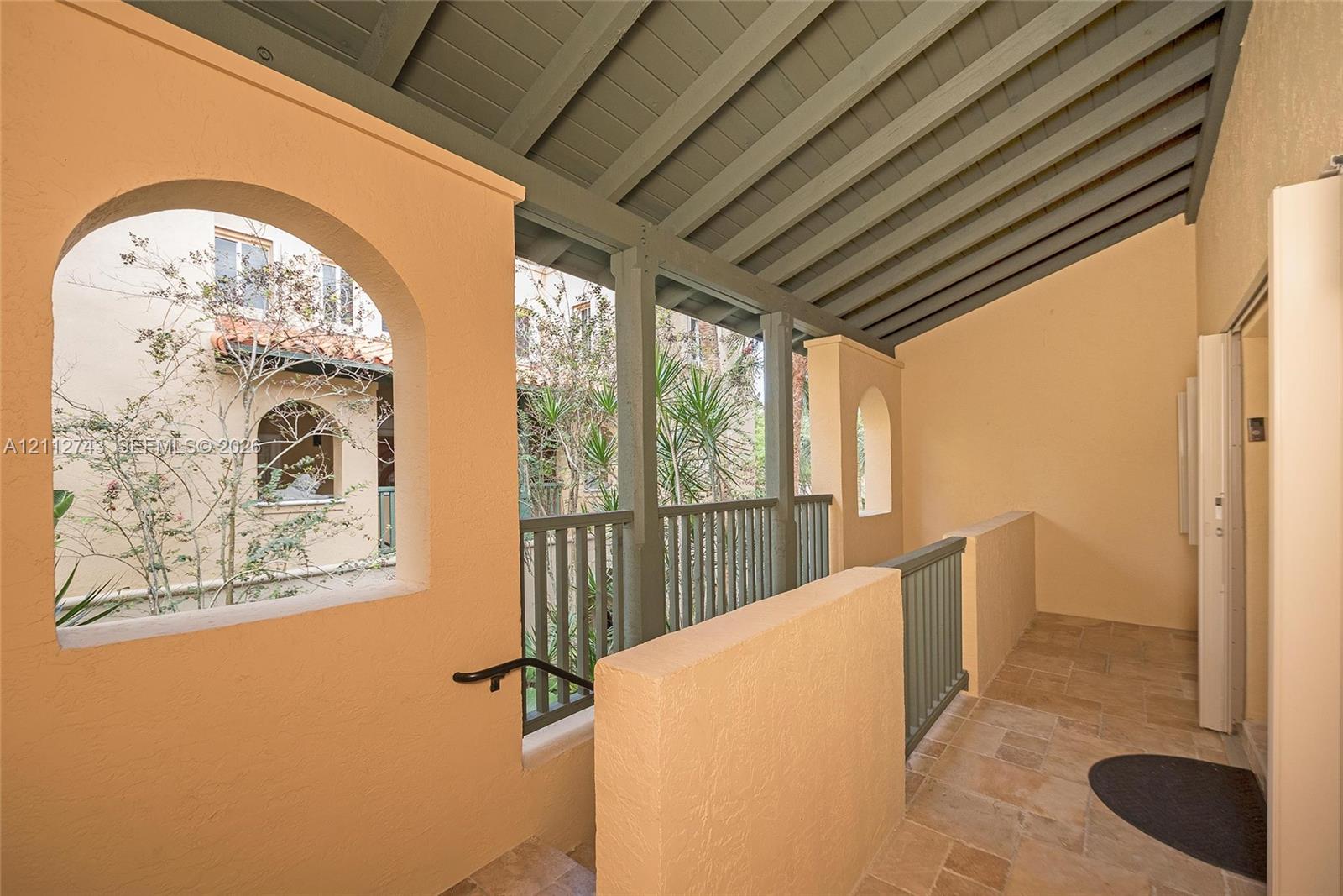Covered exterior walkway featuring arched openings, stone-tile flooring, painted wood-beam ceiling, and vertical slat railings