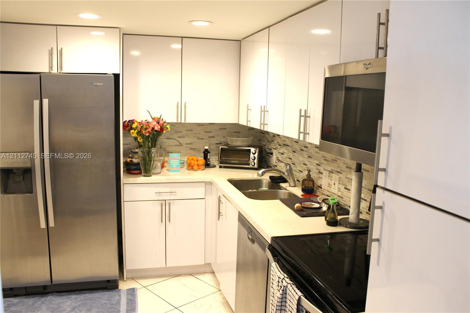 Kitchen featuring high-gloss white cabinetry, stainless steel appliances, stone-finish countertops, tile backsplash, and tile flooring