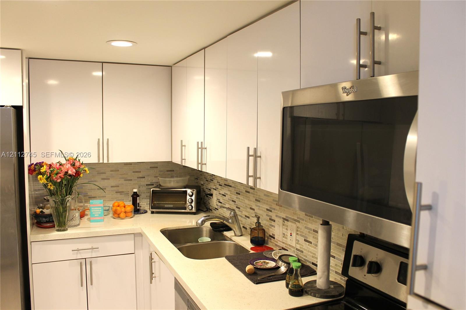 Glossy white cabinetry, solid-surface countertops, mosaic tile backsplash, recessed ceiling lighting, and stainless steel over-the-range microwave