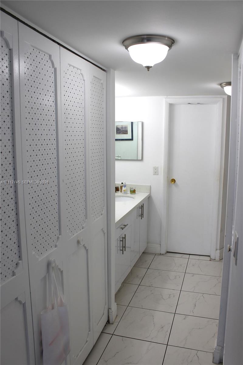 Floor-to-ceiling white cabinetry with textured detail, white vanity with rectangular under-mount sink, stone-finish tile flooring, and flush-mount lighting fixtures