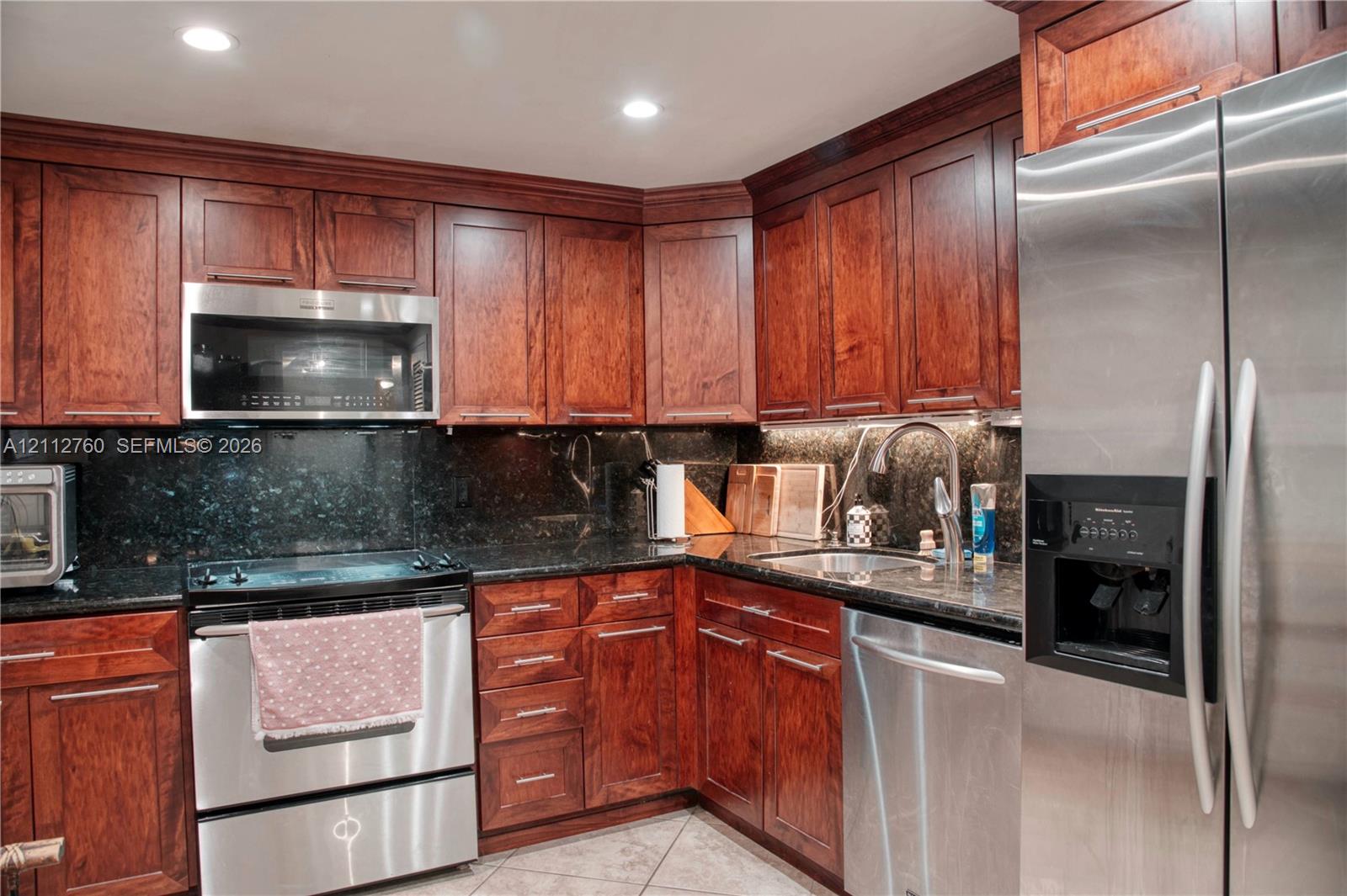Kitchen featuring cherry-finish cabinetry, dark stone countertops, stainless steel appliances, a recessed under-cabinet lighting system, and tile flooring