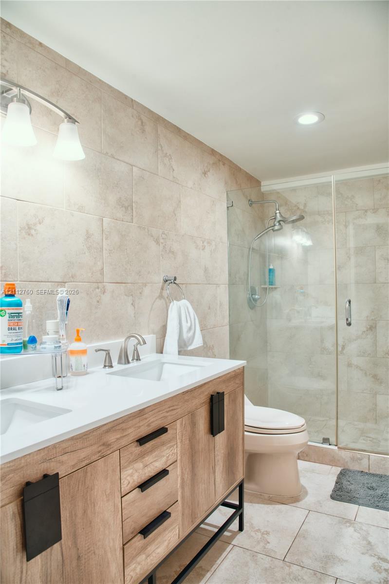 Double vanity featuring a white countertop with dual integrated sinks and a wood-finish cabinet base, complemented by a walk-in glass-enclosed shower with neutral tile surround