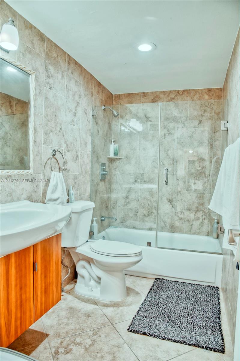 Full-wall tile surround, glass shower enclosure, wood-finish vanity cabinet, and recessed ceiling lighting