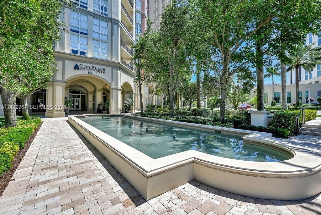 Multi-story exterior featuring a stone-clad building facade, arched entryway, paved pedestrian walkway, and a formal reflecting pool surrounded by mature landscaping