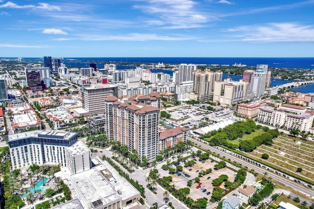 Aerial perspective featuring a high-rise residential structure, urban cityscape, dense foliage, paved parking areas, and a coastal waterway backdrop