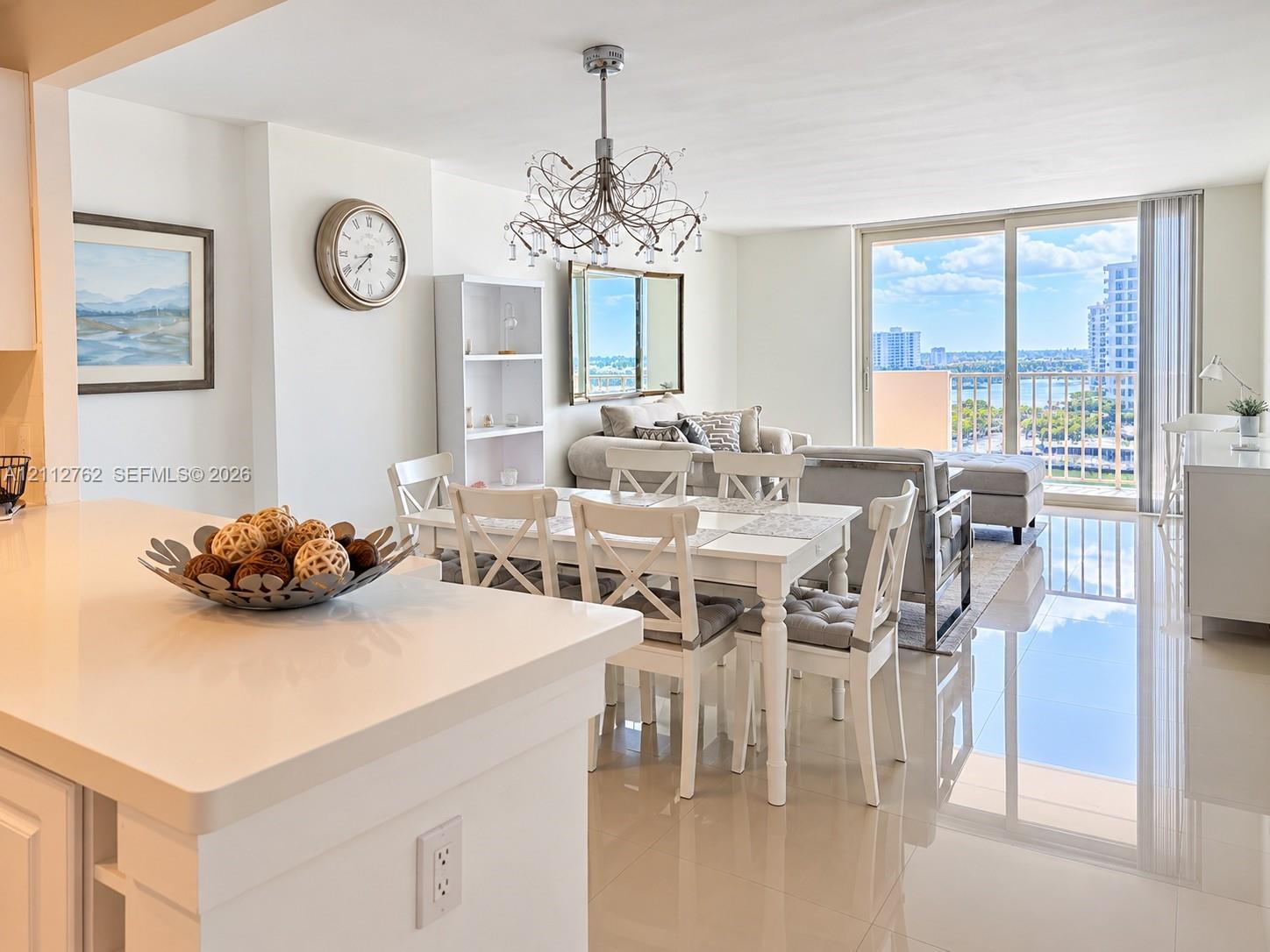 Open-concept living space featuring high-gloss tile flooring, floor-to-ceiling sliding glass doors, a decorative multi-arm chandelier, and a kitchen peninsula with solid-surface countertops
