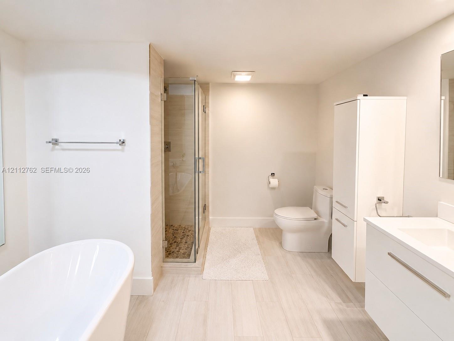 Freestanding soaking tub, glass-enclosed shower with pebble tile flooring, white floating vanity, and tall built-in storage cabinetry