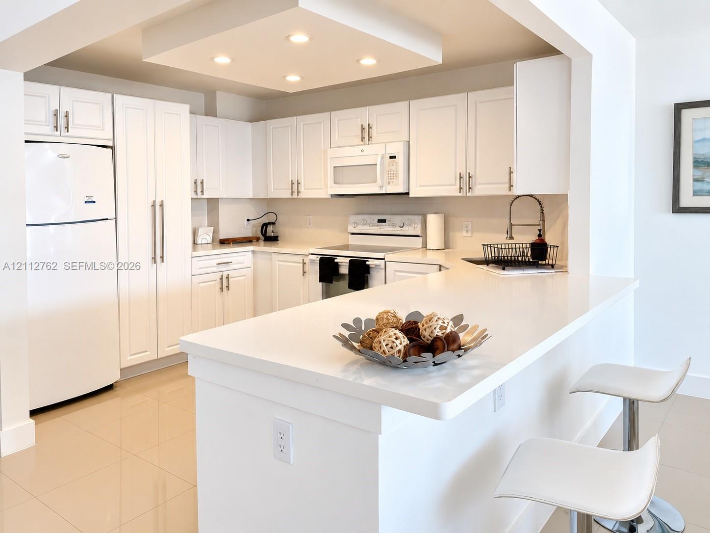 Kitchen featuring white shaker-style cabinetry, quartz countertops, a peninsula with bar seating, a recessed tray ceiling with integrated lighting, and neutral tile flooring