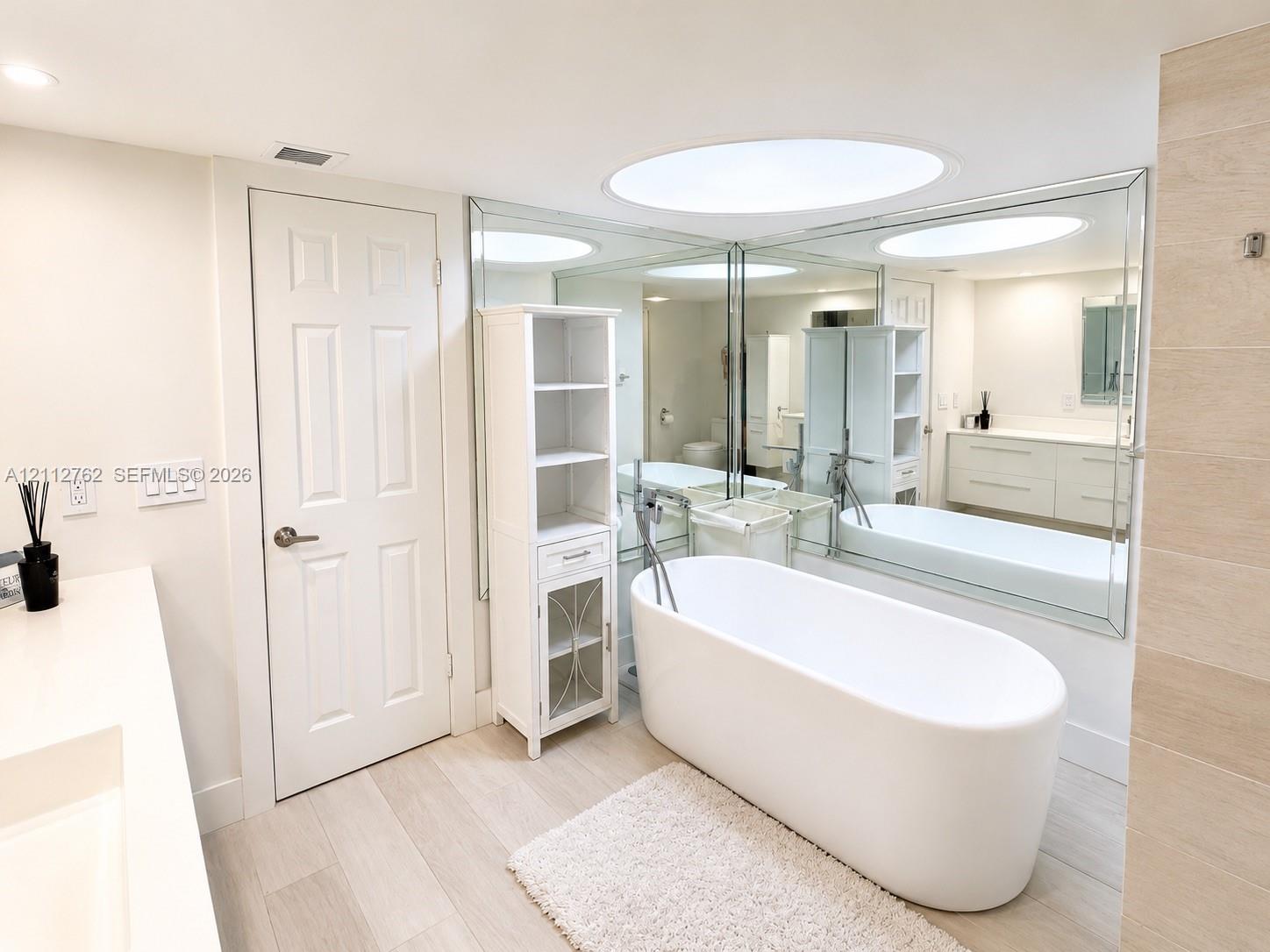 Freestanding soaking tub with a floor-mounted faucet, wall-to-wall vanity mirror, neutral-toned flooring, and built-in cabinetry