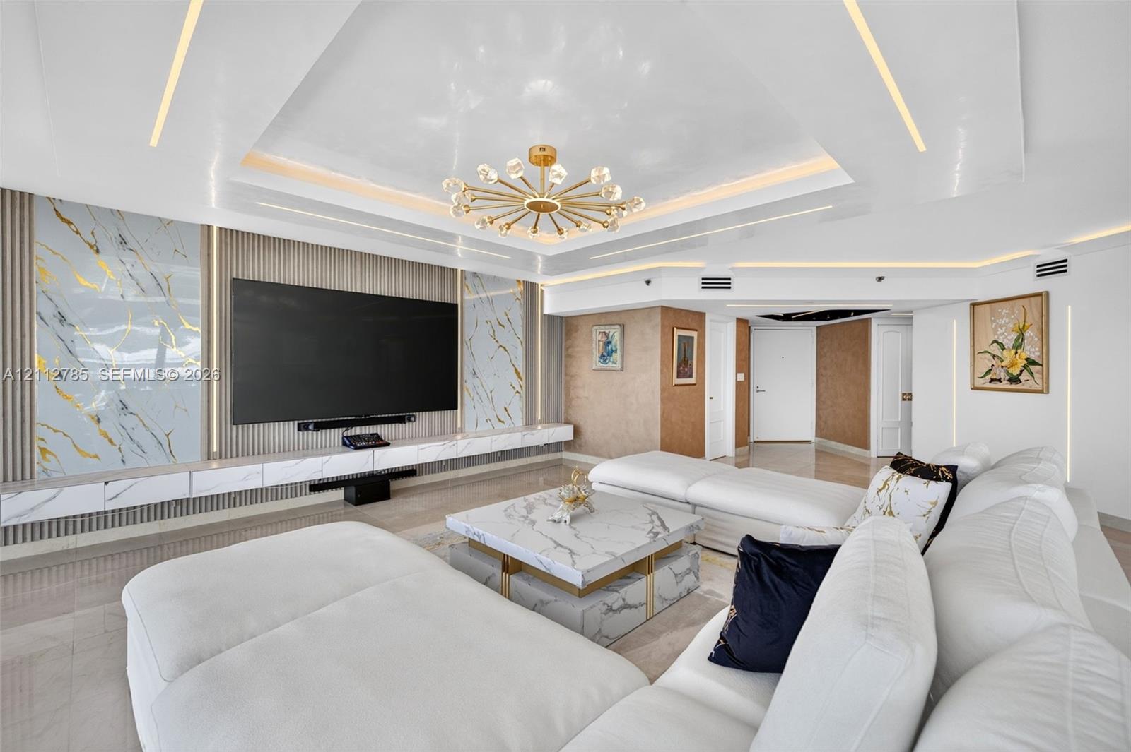 Tray ceiling featuring integrated perimeter lighting, custom stone-panel wall accents, recessed linear lighting, and a modern multi-arm chandelier Tray ceiling featuring integrated perimeter lighting, custom stone-panel wall accents, recessed linear lighting, and a modern multi-arm chandelier