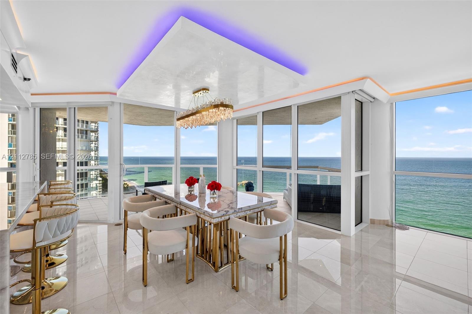 Open-concept dining area featuring floor-to-ceiling windows, polished tile flooring, a multi-tiered crystal chandelier, and a recessed ceiling cove with integrated lighting