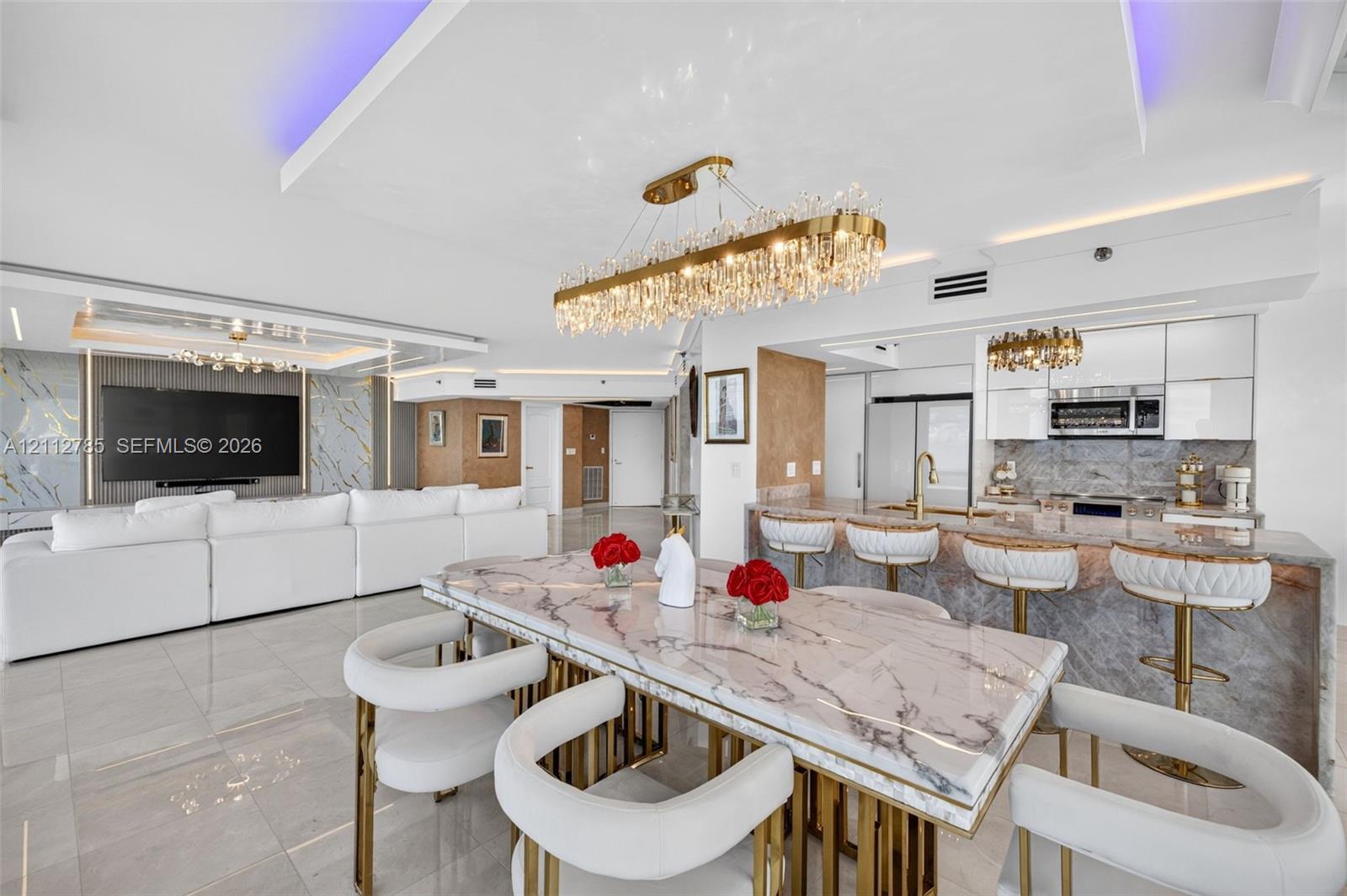 Open-concept living space featuring high-gloss tile flooring, stone-surfaced island and dining table, gold-toned crystal chandeliers, and recessed tray ceilings with integrated ambient lighting