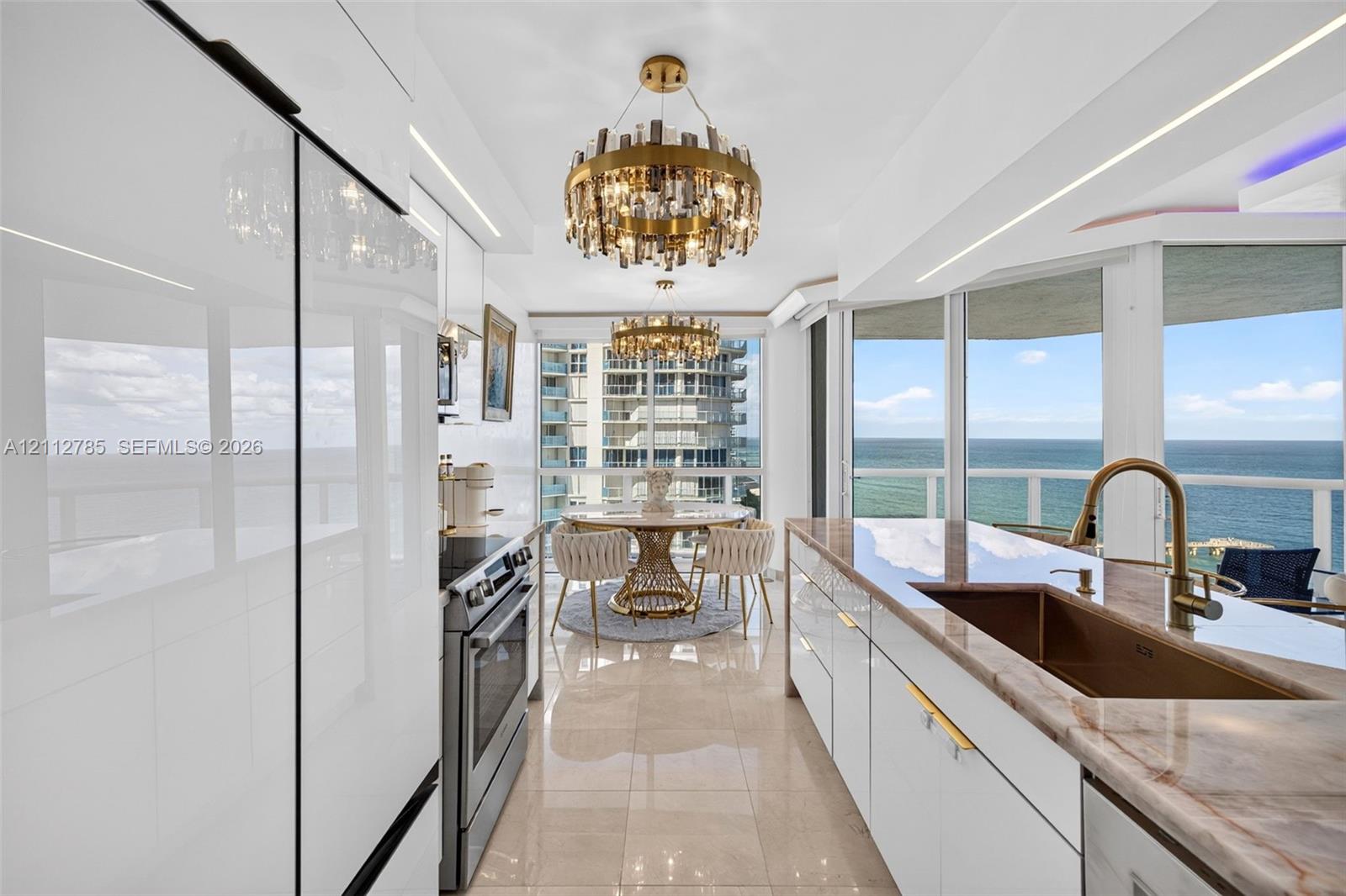 Contemporary kitchen featuring high-gloss white cabinetry, stone-surface countertops, integrated stainless steel appliances, tiered crystal chandeliers, and floor-to-ceiling windows with coastal scenery