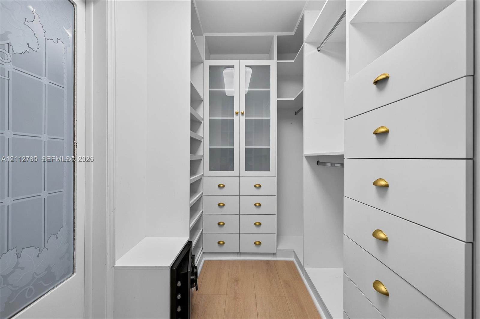 Custom walk-in closet featuring white built-in cabinetry, glass-front upper storage, soft-gold tone hardware, integrated drawer banks, and wood-finish flooring