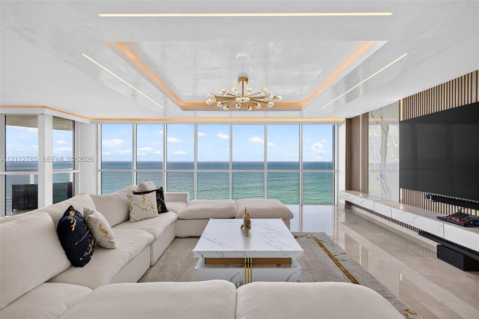 Floor-to-ceiling windows featuring ocean scenery, tray ceiling with recessed cove lighting, central sputnik-style chandelier, and custom built-in media wall with stone cladding and vertical wood slat detailing Floor-to-ceiling windows featuring ocean scenery, tray ceiling with recessed cove lighting, central sputnik-style chandelier, and custom built-in media wall with stone cladding and vertical wood slat detailing