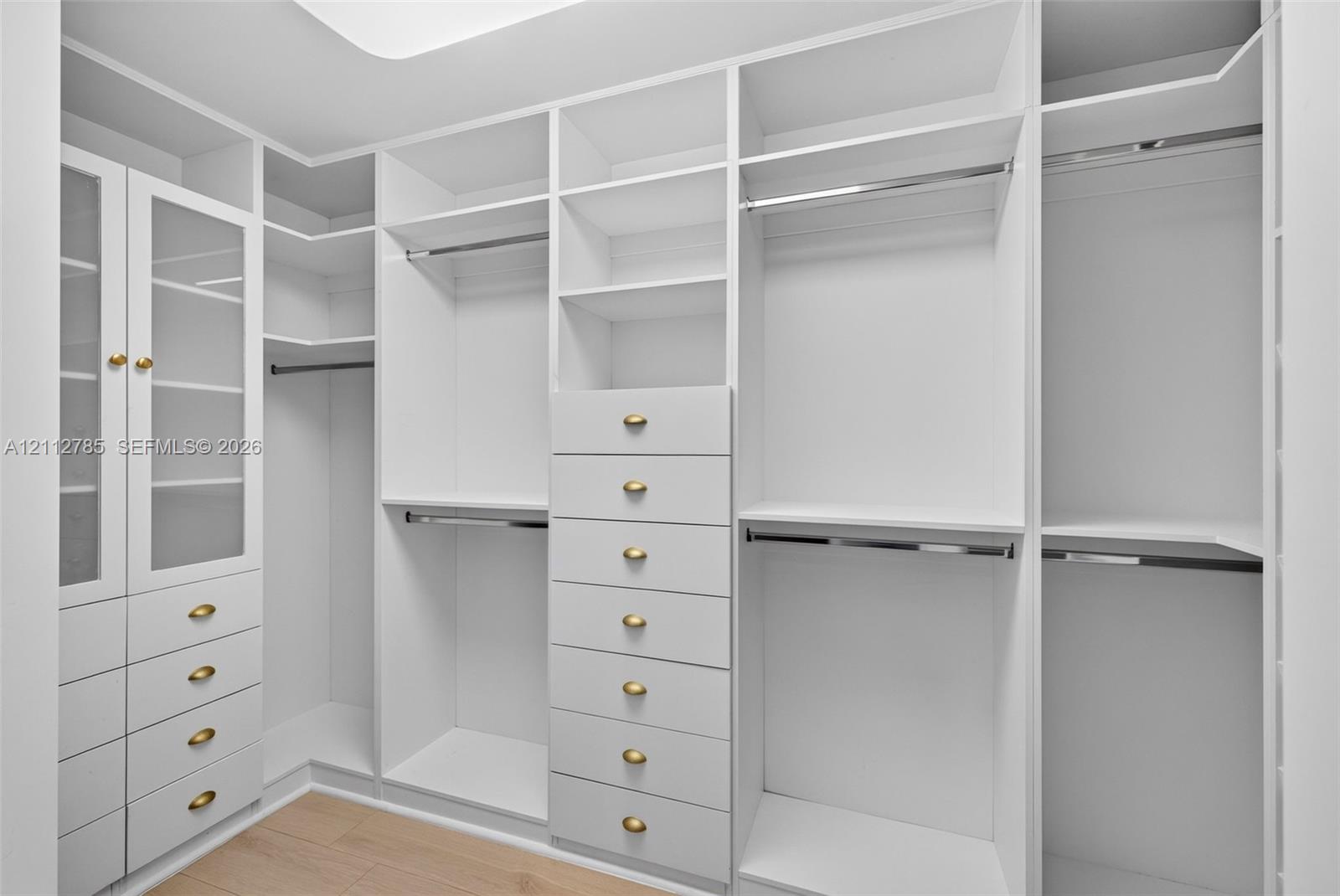 Custom walk-in closet featuring white built-in cabinetry, glass-front upper storage, multiple hanging rods, and integrated drawers with metallic hardware finished with wood-finish flooring